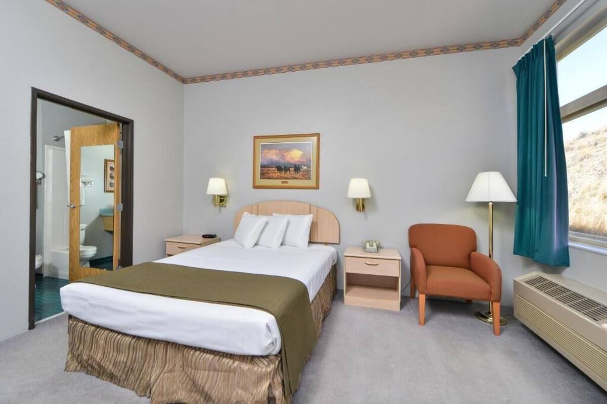 A hotel room with a bed, two bedside tables, a chair, a window with curtains, a wall-mounted picture, and a radiator.