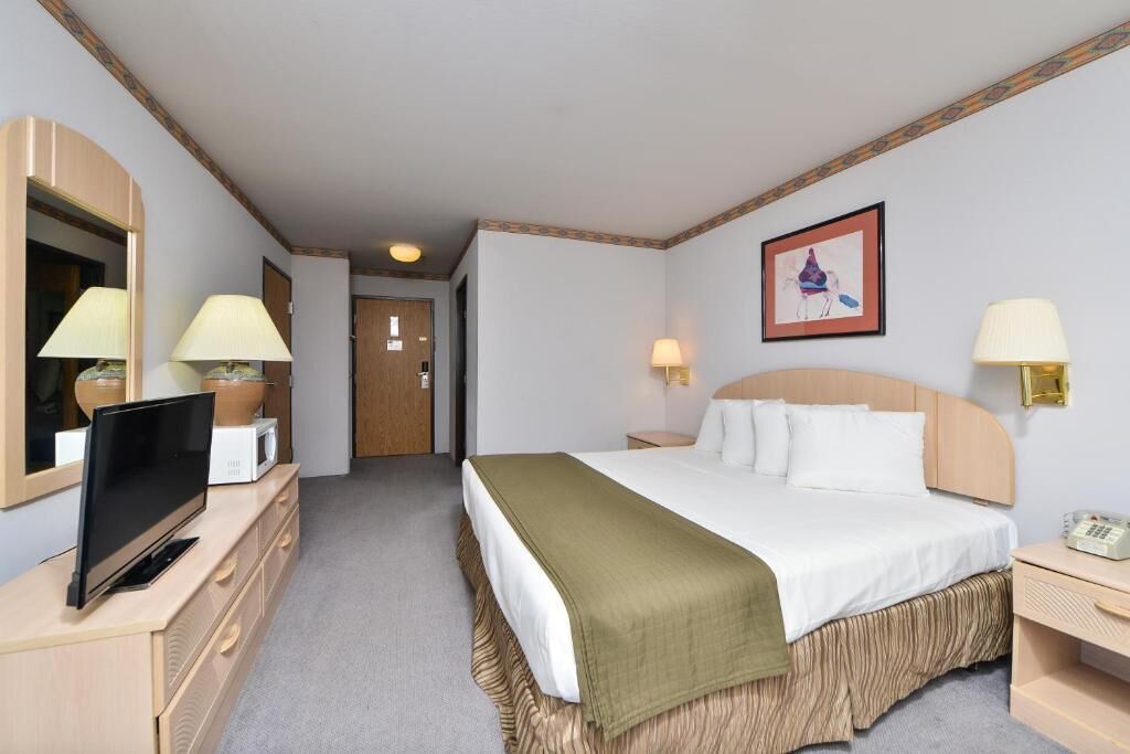Una habitación de hotel con cama, lámparas de noche, televisor en una cómoda, un cuadro en la pared y una puerta que da a otra habitación.