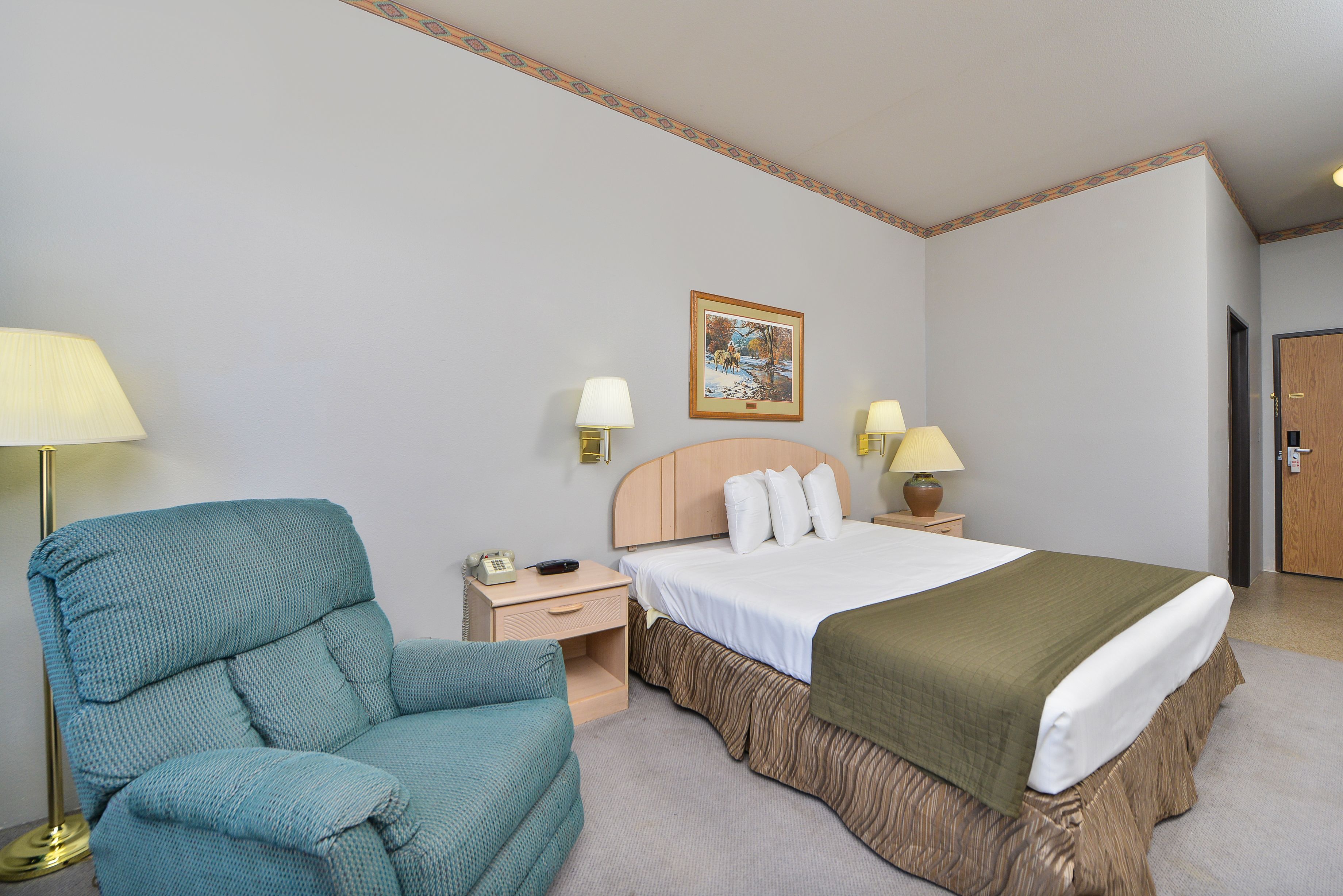 A hotel room with a bed, a nightstand, a lamp, a chair, and a painting on the wall.