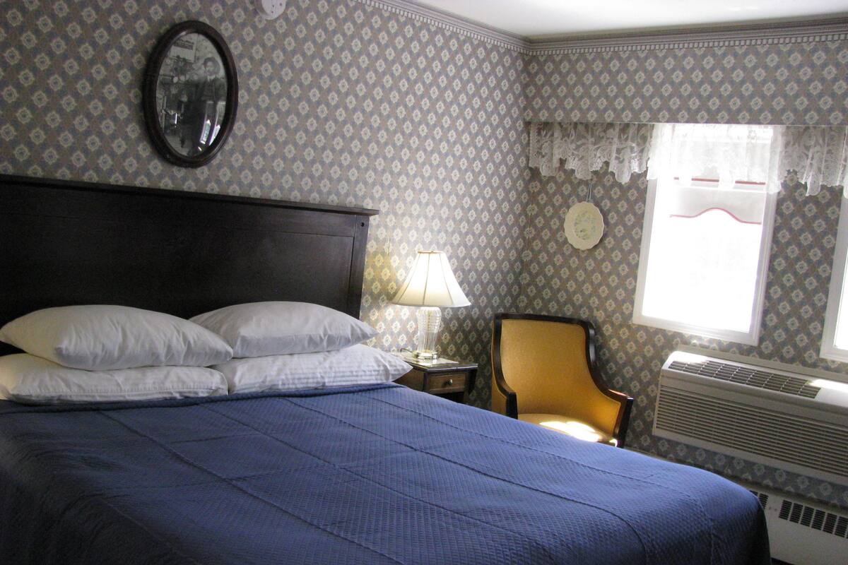 A neatly made bed with white pillows, a bedside table with a lamp, a chair, and a window with a lace curtain.