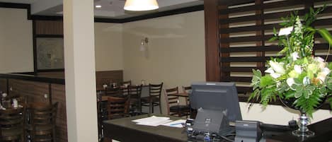 A hotel reception area with a wooden reception desk, a computer, and a telephone. There are several wooden chairs and tables in the background.