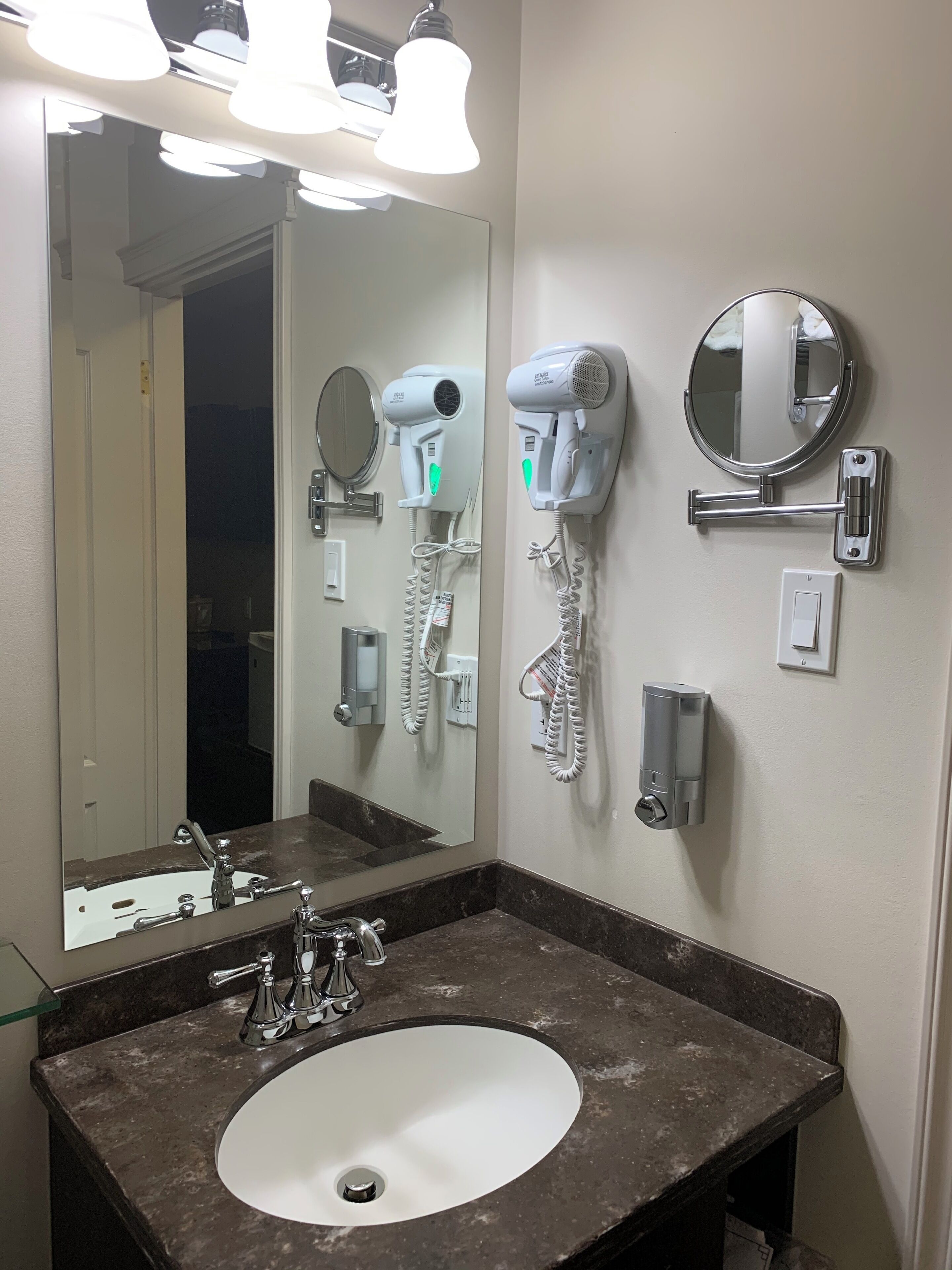 Un baño con lavamanos, espejo, secadores de pelo y dispensador de toallas.