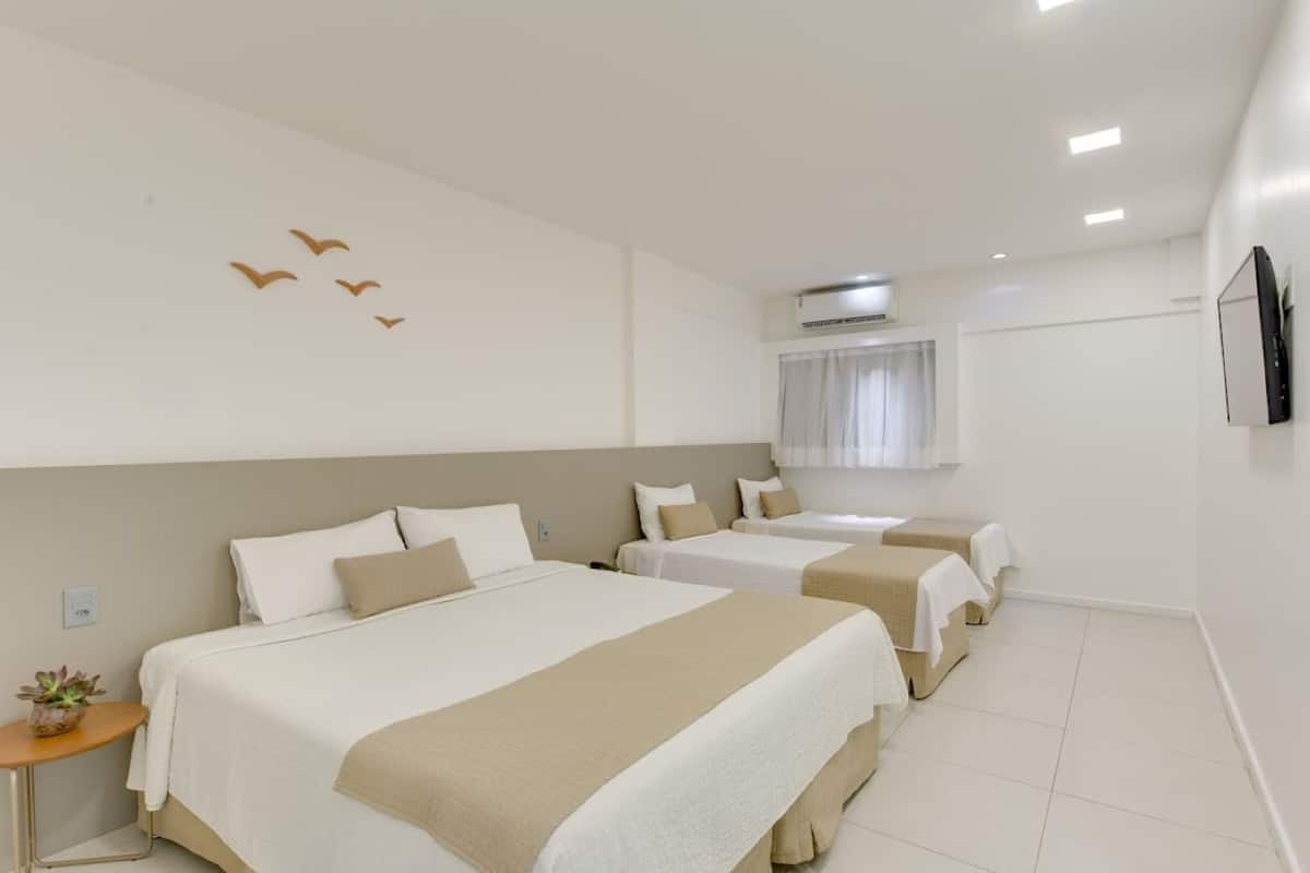 A hotel room with three beds, a small table with a plant, an air conditioning unit, and a TV mounted on the wall.