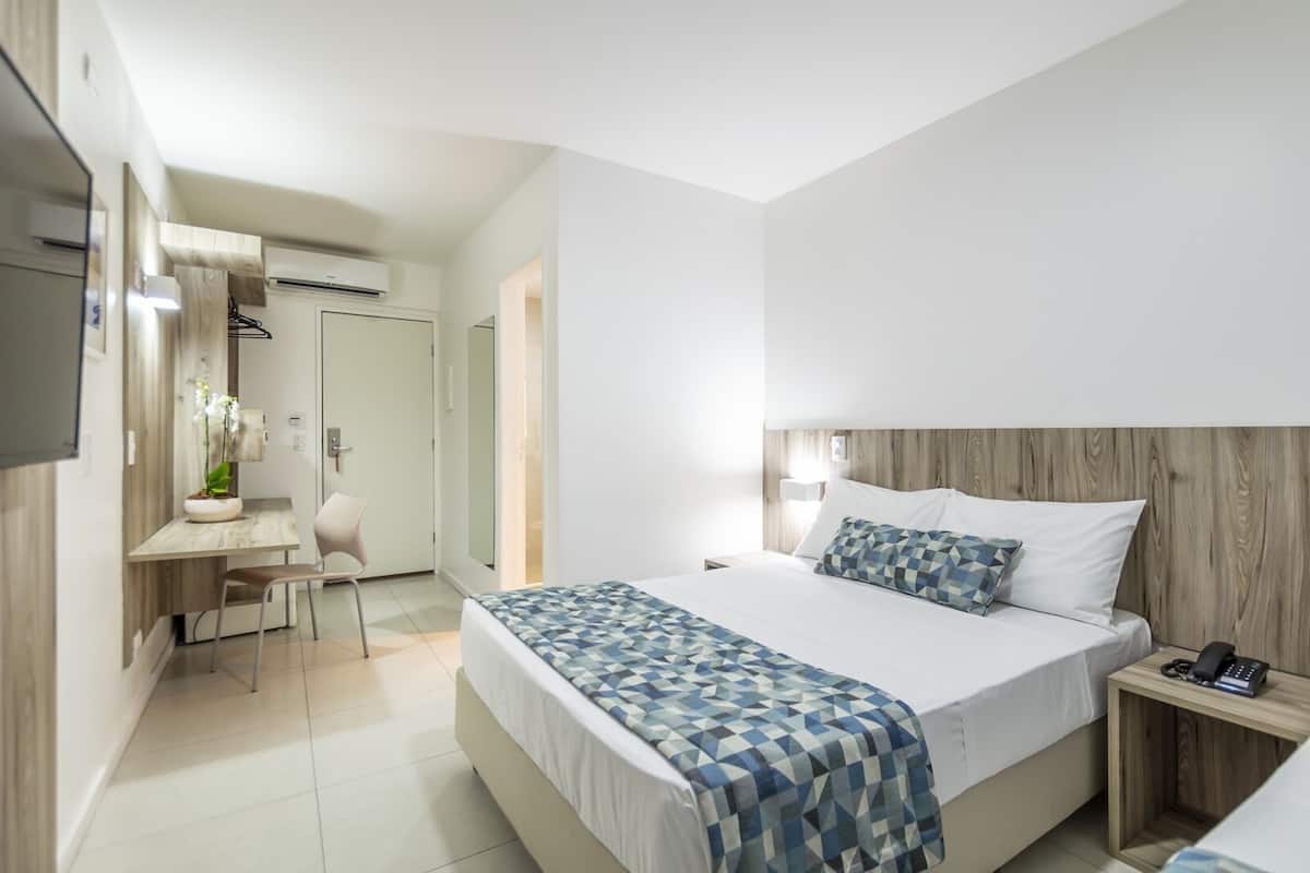 A modern hotel room with a bed, bedside table, desk, chair, and a TV.