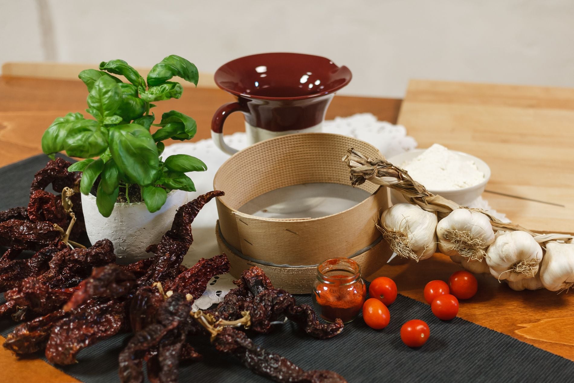 Getrocknete rote Chilischoten, frische Tomaten, Knoblauch, eine Wasserschüssel, ein Topf mit Basilikum und eine rote Tasse auf einem Holztisch.