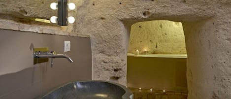 Un baño con lavabo de piedra, espejo con iluminación y una bañera en un ambiente que recuerda a una cueva.