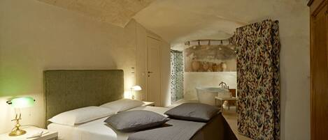 A bedroom with a bed, bedside lamps, a nightstand, a bathroom with a sink and mirror, and a shower area.