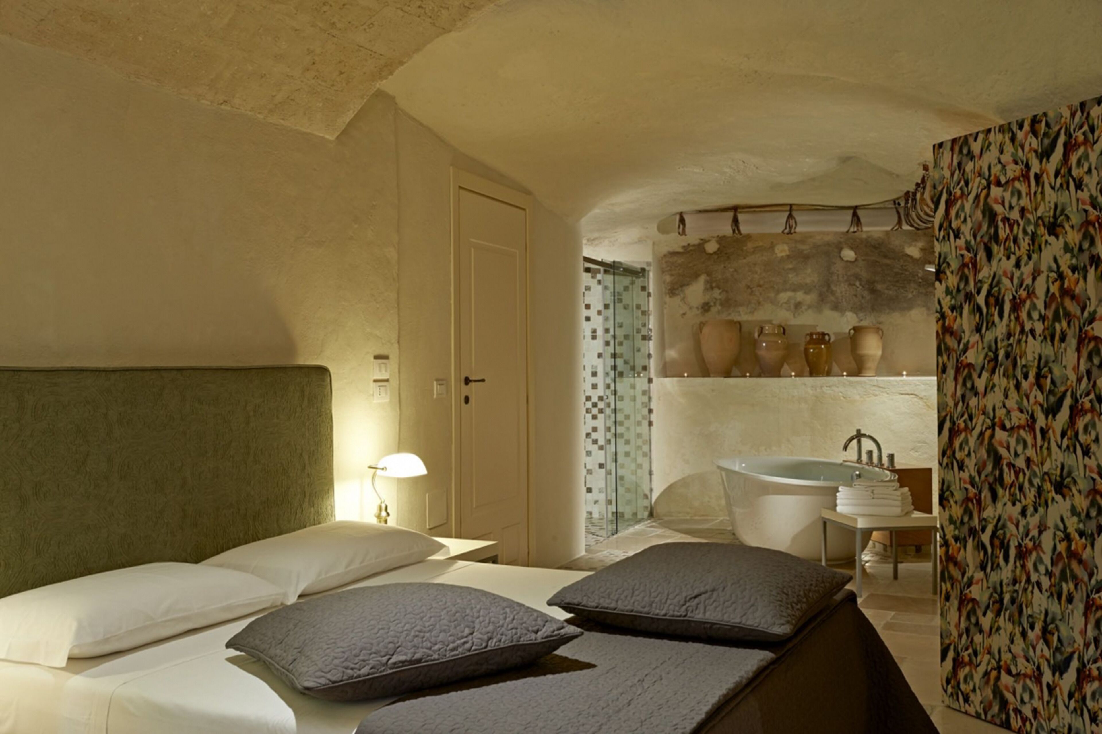 A bedroom with a bed, pillows, a nightstand, a lamp, a bathroom with a sink and a bathtub, and a patterned wall.
