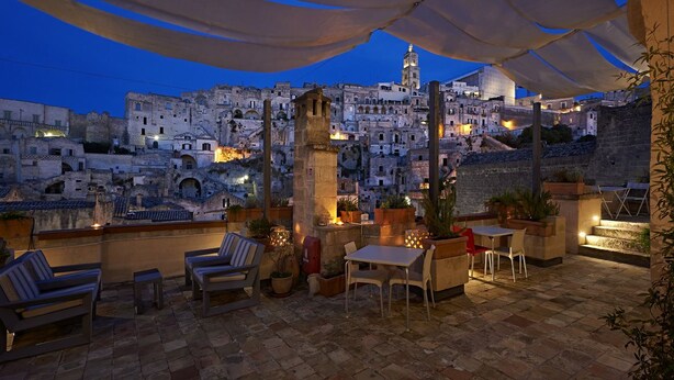 An outdoor terrace with seating, potted plants, and a view of a historic cityscape at dusk. - Fra i Sassi - Residence (Matera)