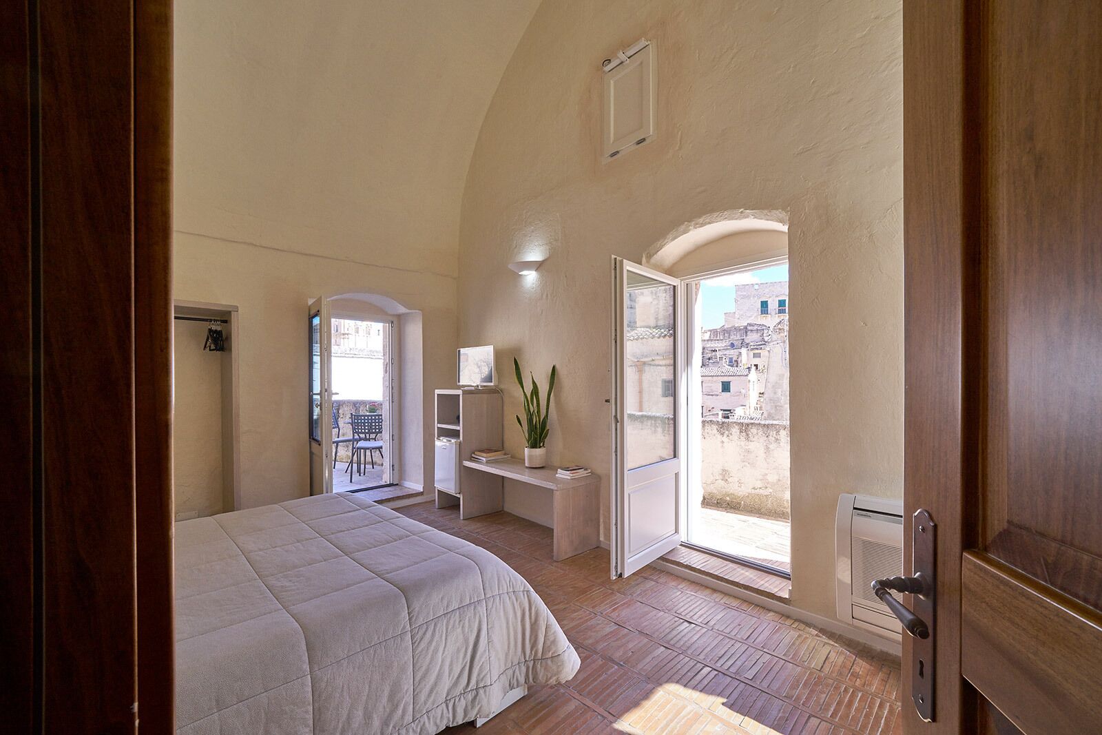 A bedroom with a bed, a desk, a chair, and a view of the city through an open door.