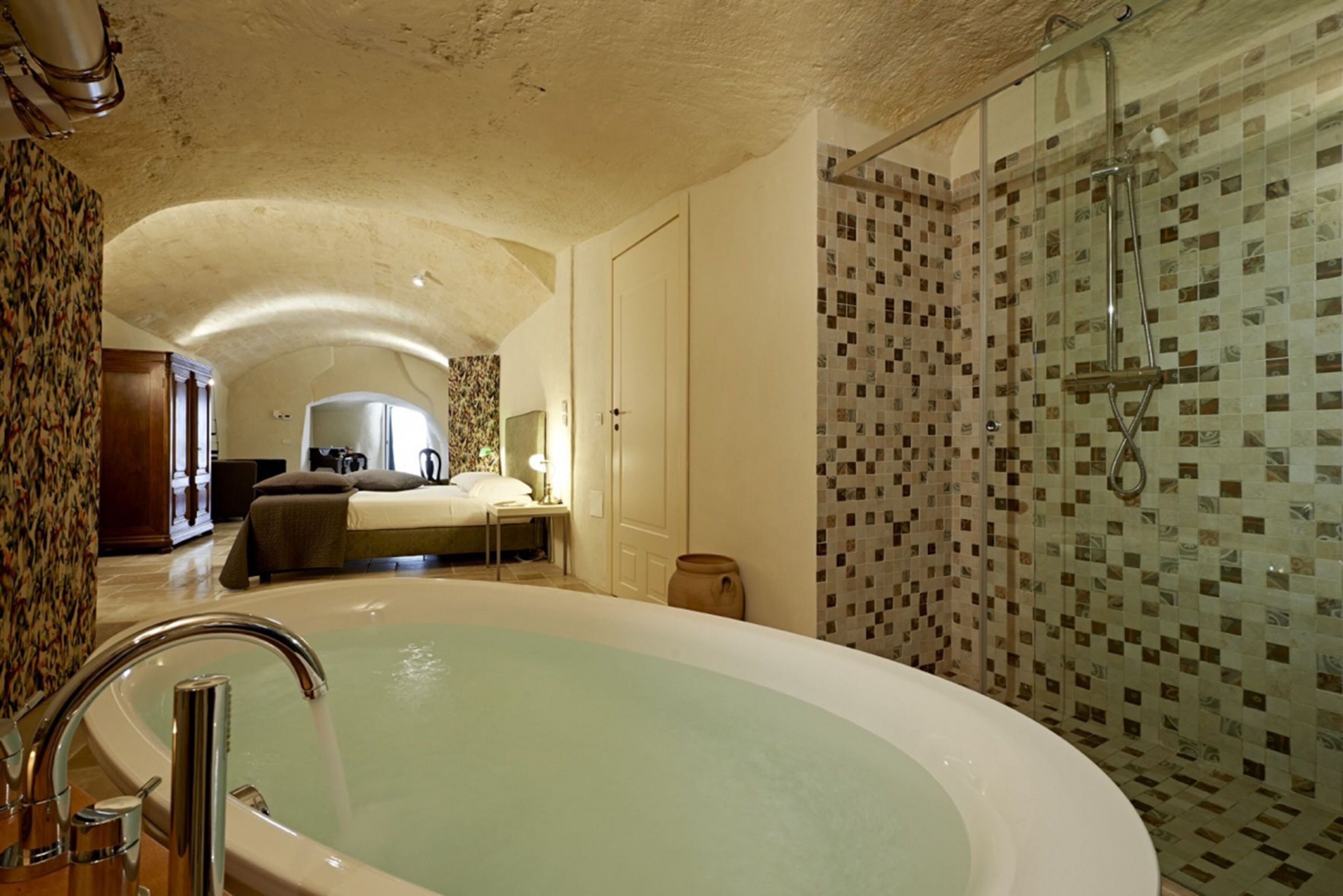 A spacious bathroom with a large bathtub, a shower area with mosaic tiles, and a visible bedroom with a bed and nightstand.