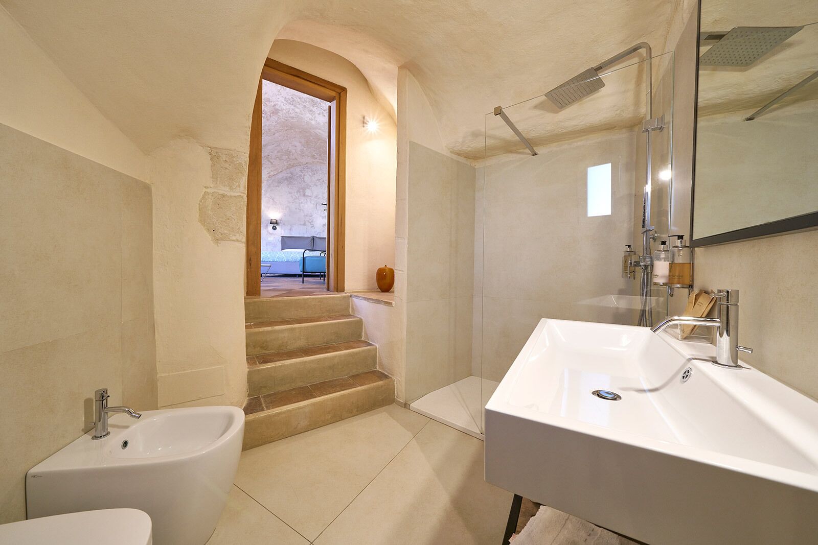 A bathroom with a large sink, a bidet, and a shower. There is a staircase leading to a bedroom.