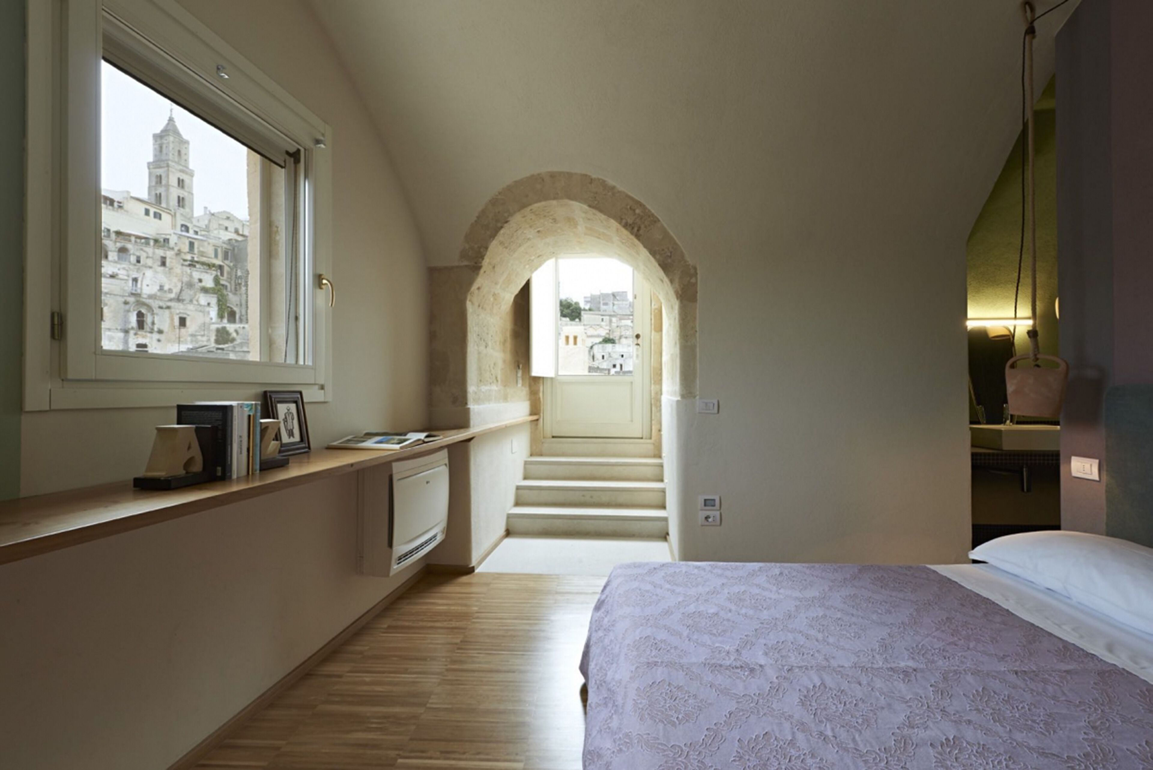Un dormitorio con una cama grande, un ventanal con vista a edificios, piso de madera y una escalera que sube a un nivel superior.