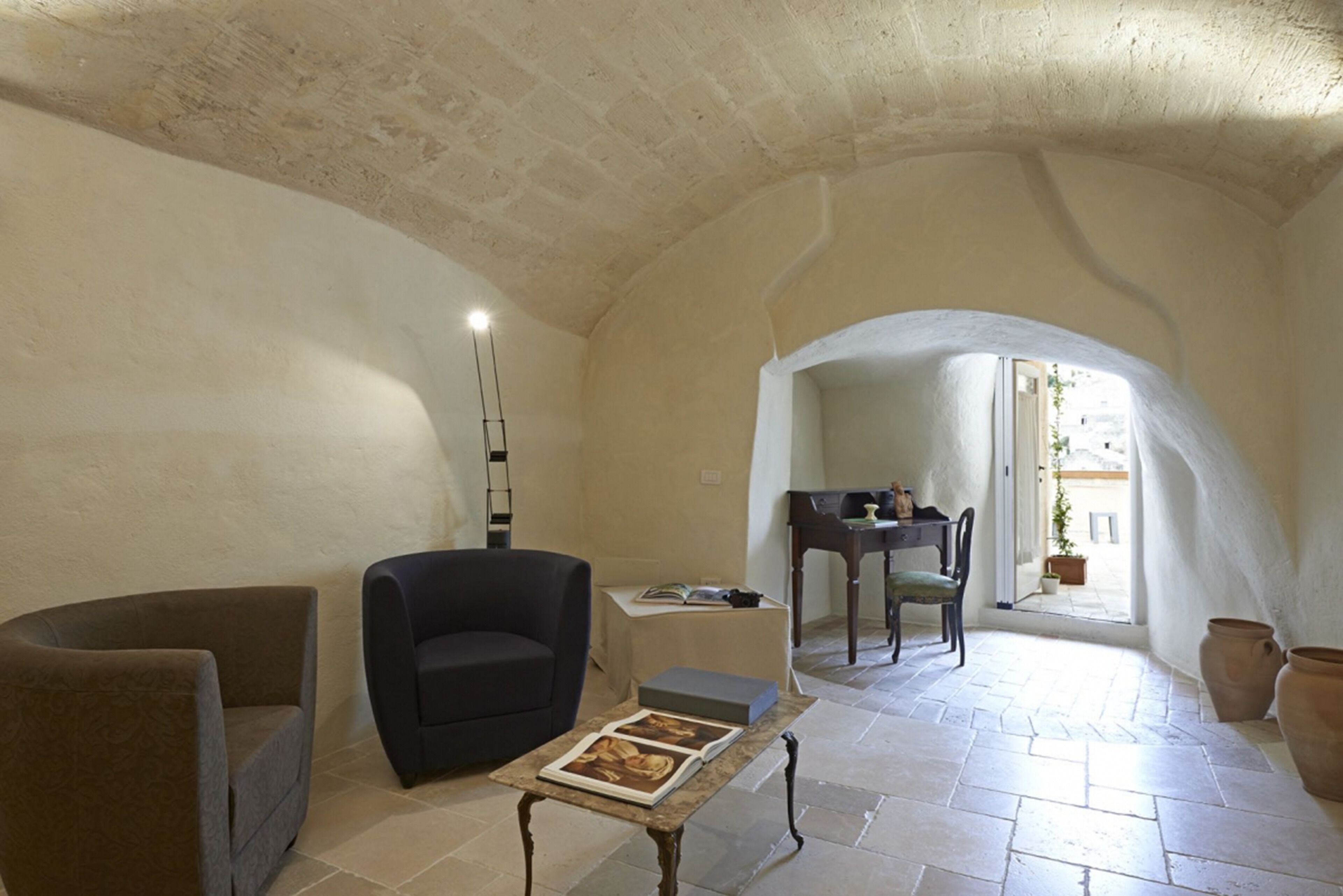 Un salón espacioso con techo alto, un sillón oscuro, una mesita con un libro y vistas al exterior a través de un arco.