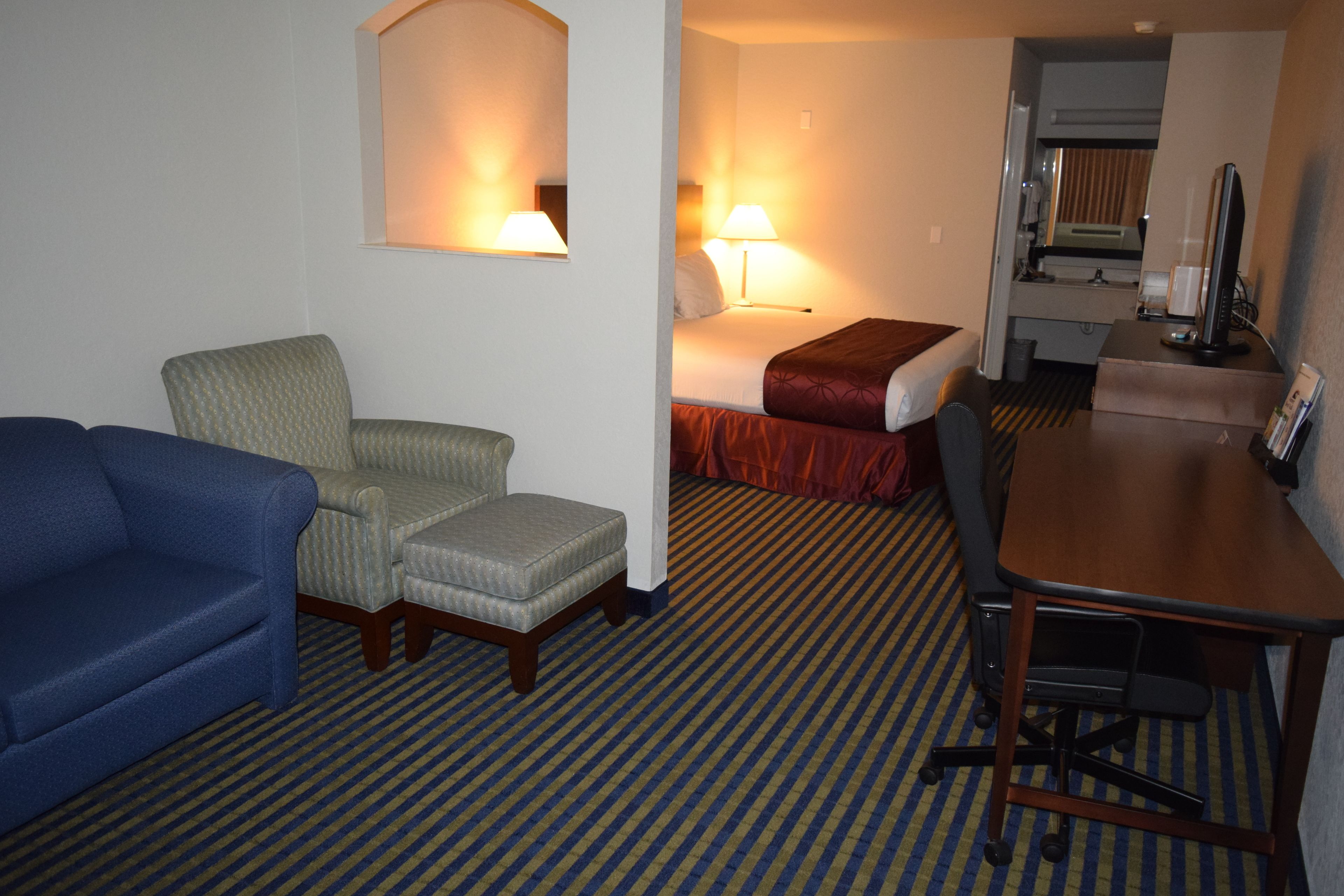 Habitación de hotel con un sofá azul, un sillón pequeño, una cama con colcha roja, un escritorio con computadora y un espejo.