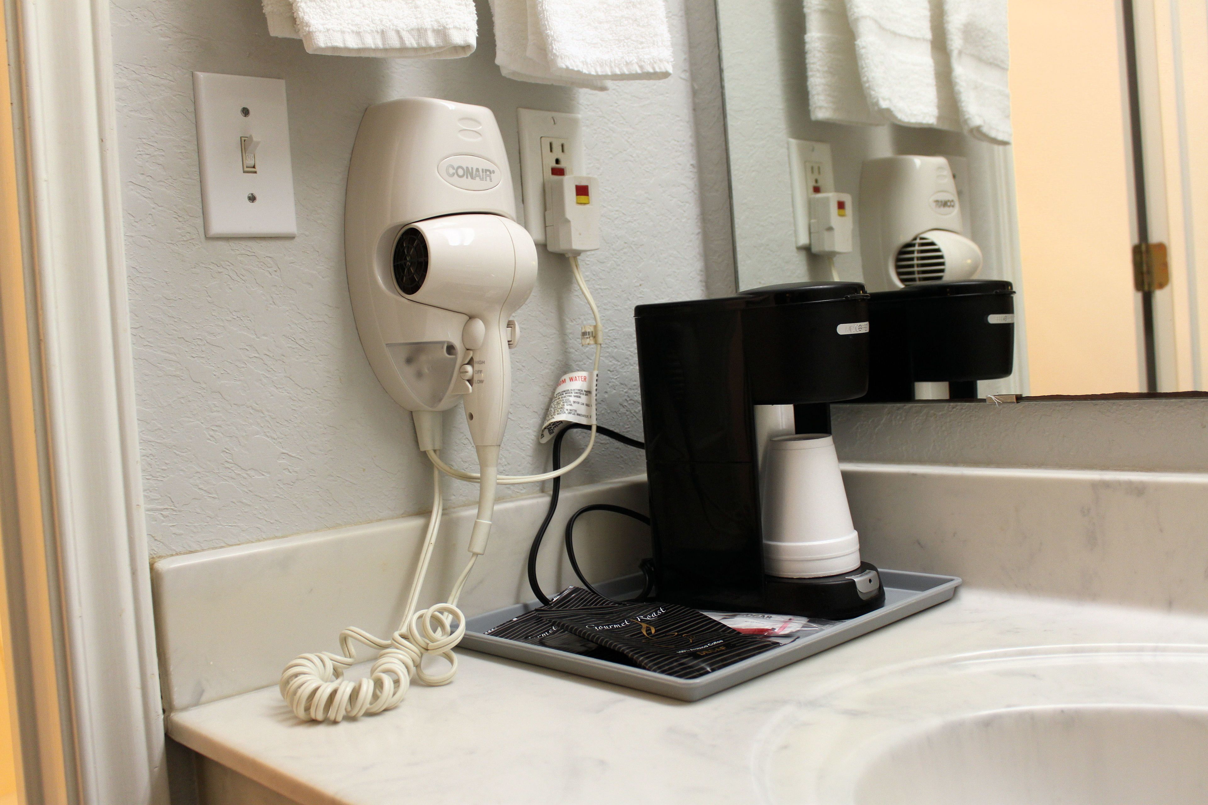 A hairdryer and coffee maker on a bathroom counter, with towels hanging on a rack in the background.
