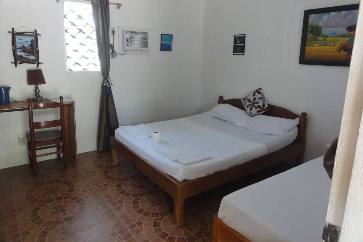 A single bed with a wooden frame, a desk with a chair, an air conditioning unit, and a window with curtains.