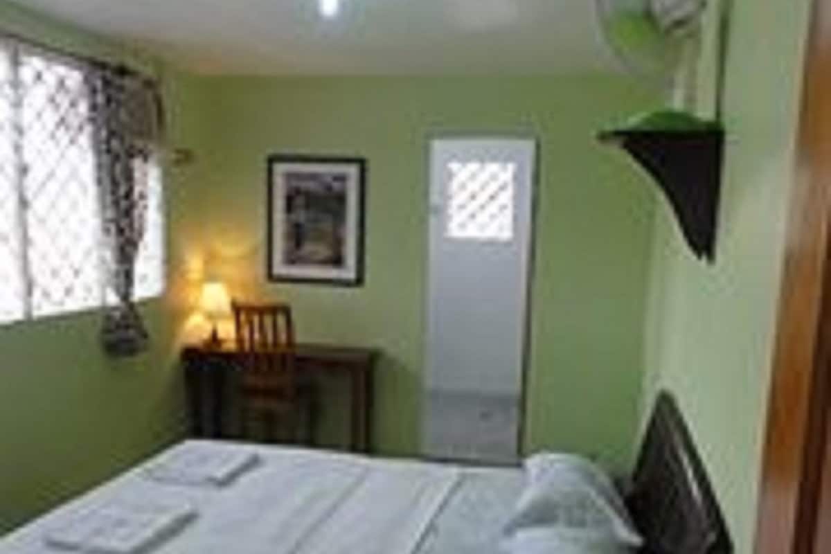 A small, single-bed room with a bed, a chair, a desk, a wall-mounted fan, and a door.