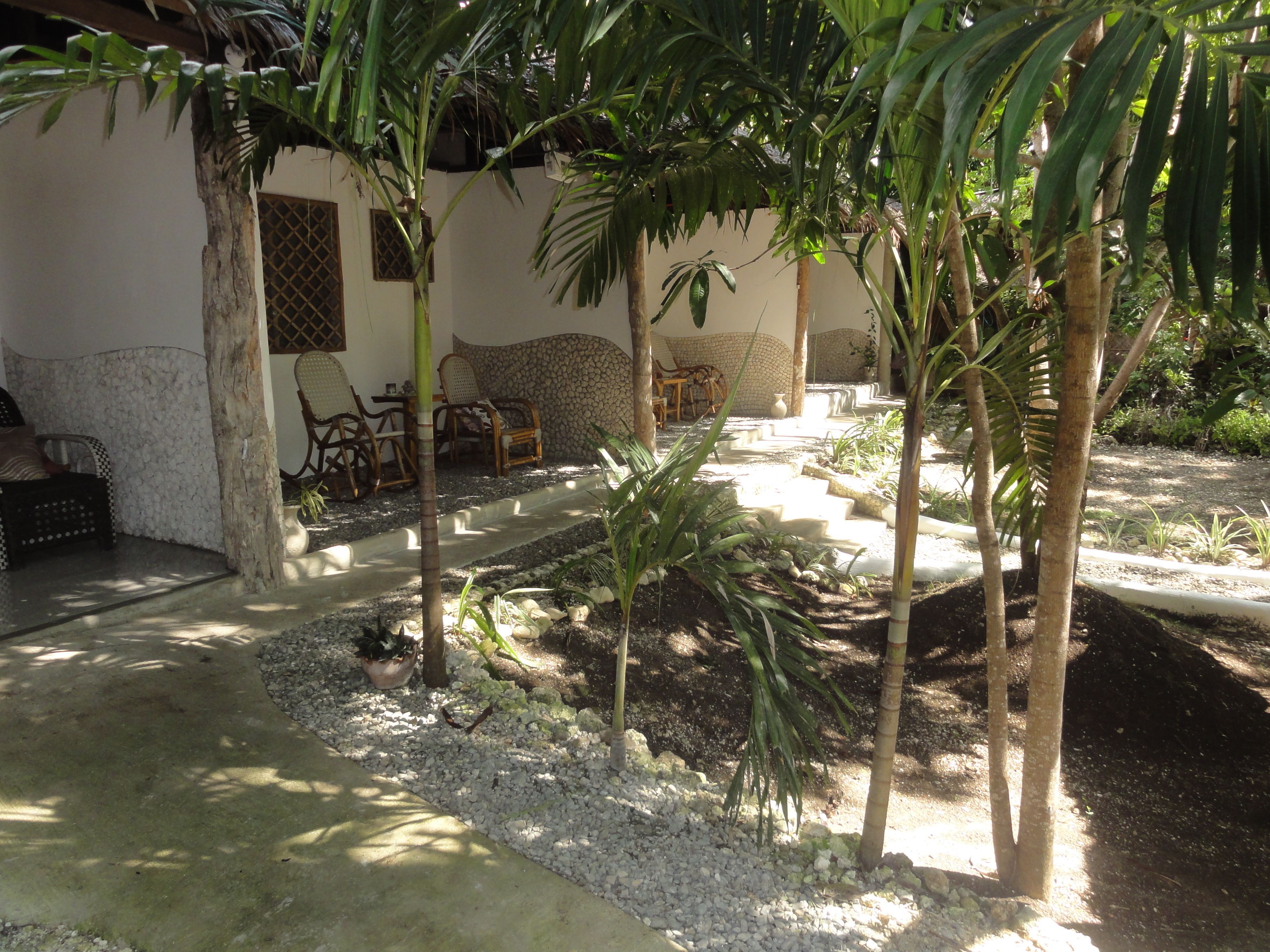 Eine Terrasse mit Sitzgelegenheit, ein kleiner Garten mit Palmen und eine Steinmauer.