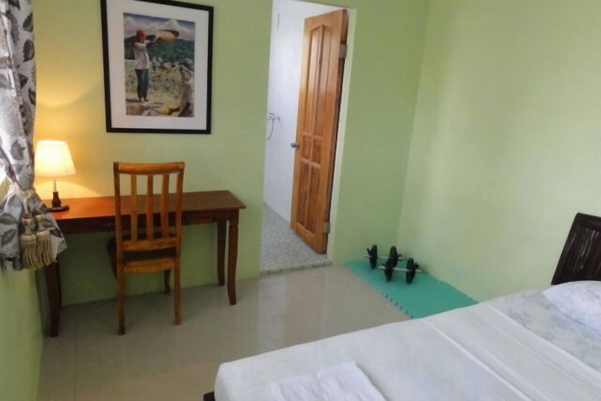 A room with a bed, a desk with a chair, a framed picture on the wall, and a door leading to another room.