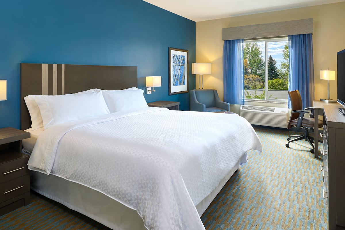 Quarto de hotel com uma cama grande, duas mesas de cabeceira com luminárias, uma escrivaninha com cadeira e uma janela ampla com cortinas.
