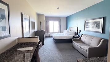 A hotel room with a bed, a sofa, a desk with a chair, a large window, and a bathroom.