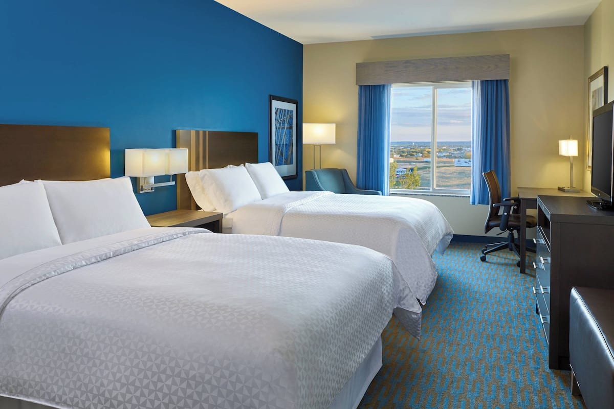 Quarto de hotel com duas camas, uma escrivaninha, uma cadeira, televisão e janela com vista.