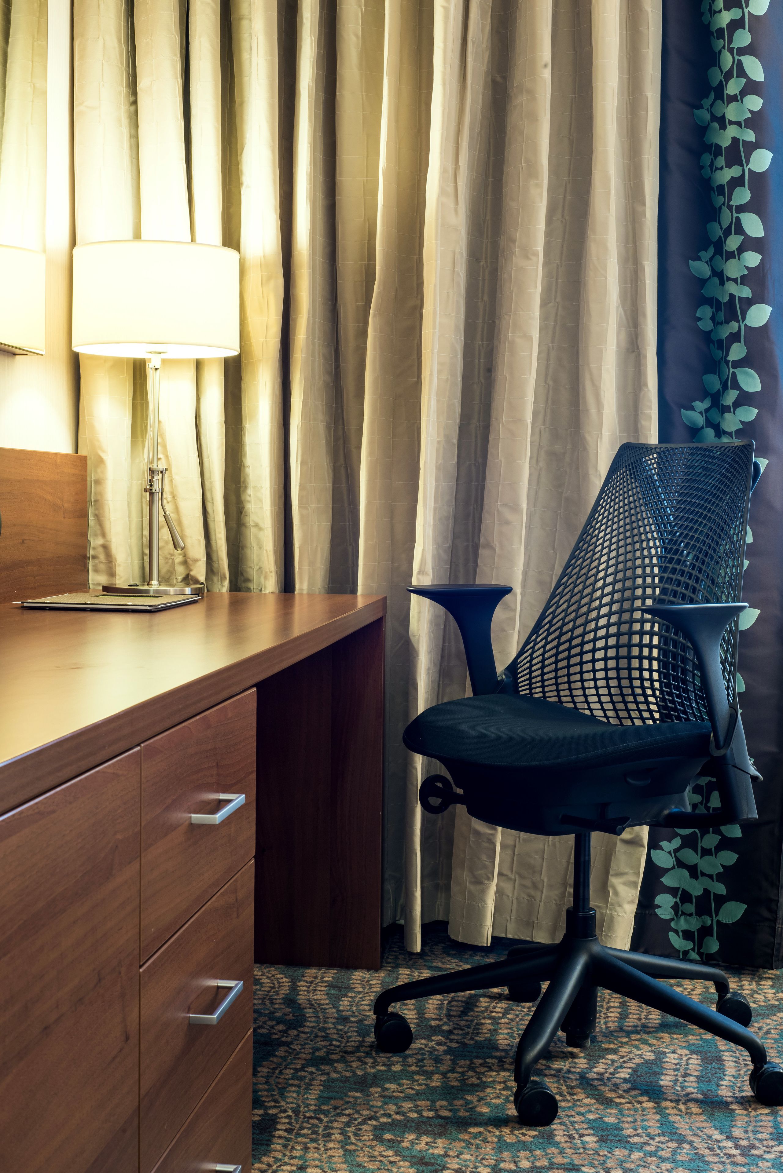 A wooden desk with a lamp, a mesh office chair, and a patterned carpet.