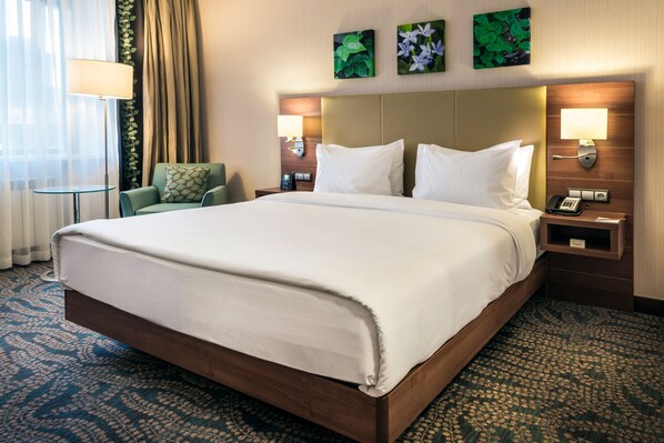 A large bed with a wooden frame, a green armchair, and a small table with a lamp. There are two wall-mounted lamps and a telephone on the nightstand. - Hilton Garden Inn Astana (Astana)