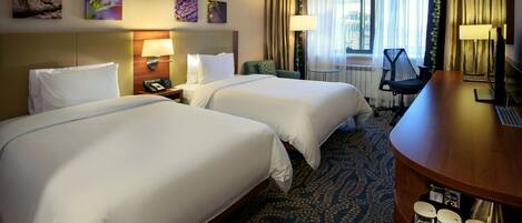 A hotel room with two beds, a desk, a chair, a nightstand, and a window with curtains.