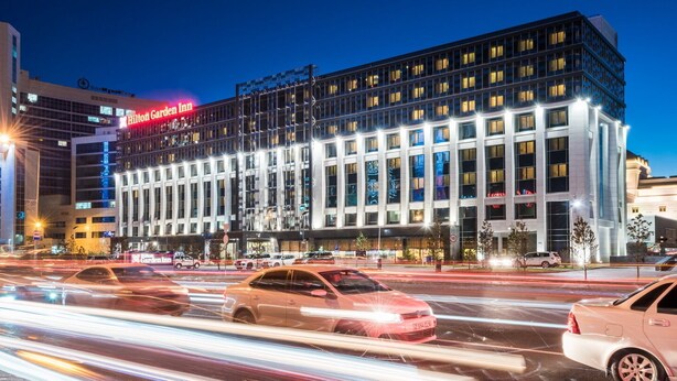 A modern hotel with a prominent Hilton Garden Inn sign, multiple floors, and a busy street with light trails indicating traffic. - Hilton Garden Inn Astana (Astana)