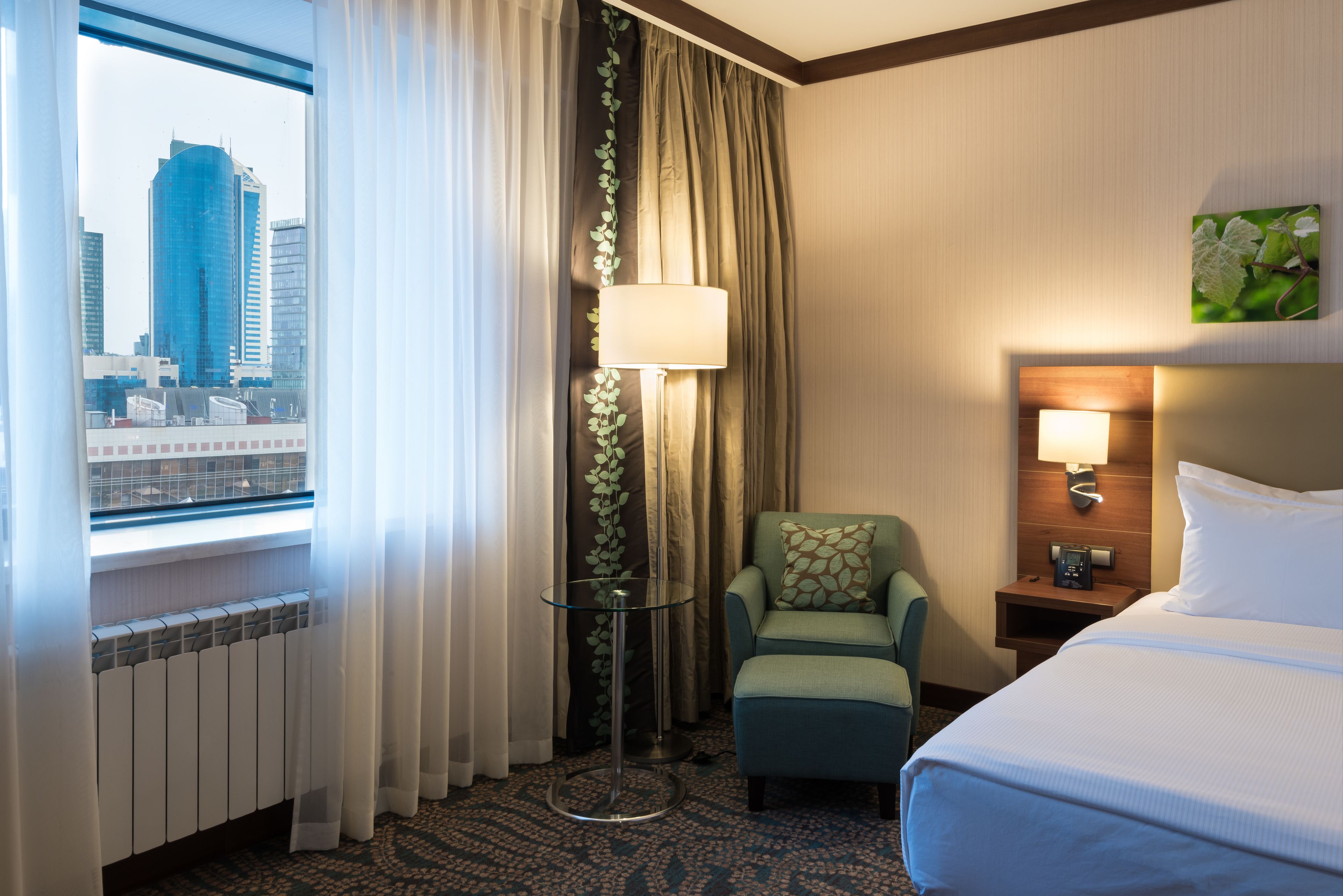 Ein Hotelzimmer mit Bett, Sessel, Tisch, Lampe, Nachttisch, Blick auf eine Stadtlandschaft durch ein Fenster und einer Heizung.