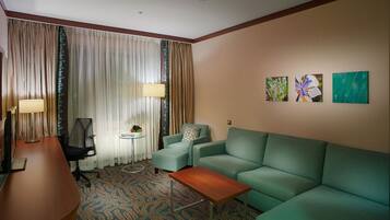 A hotel room with a green sofa, a wooden desk, a chair, a small table, and a lamp. There are curtains and a window with a view.