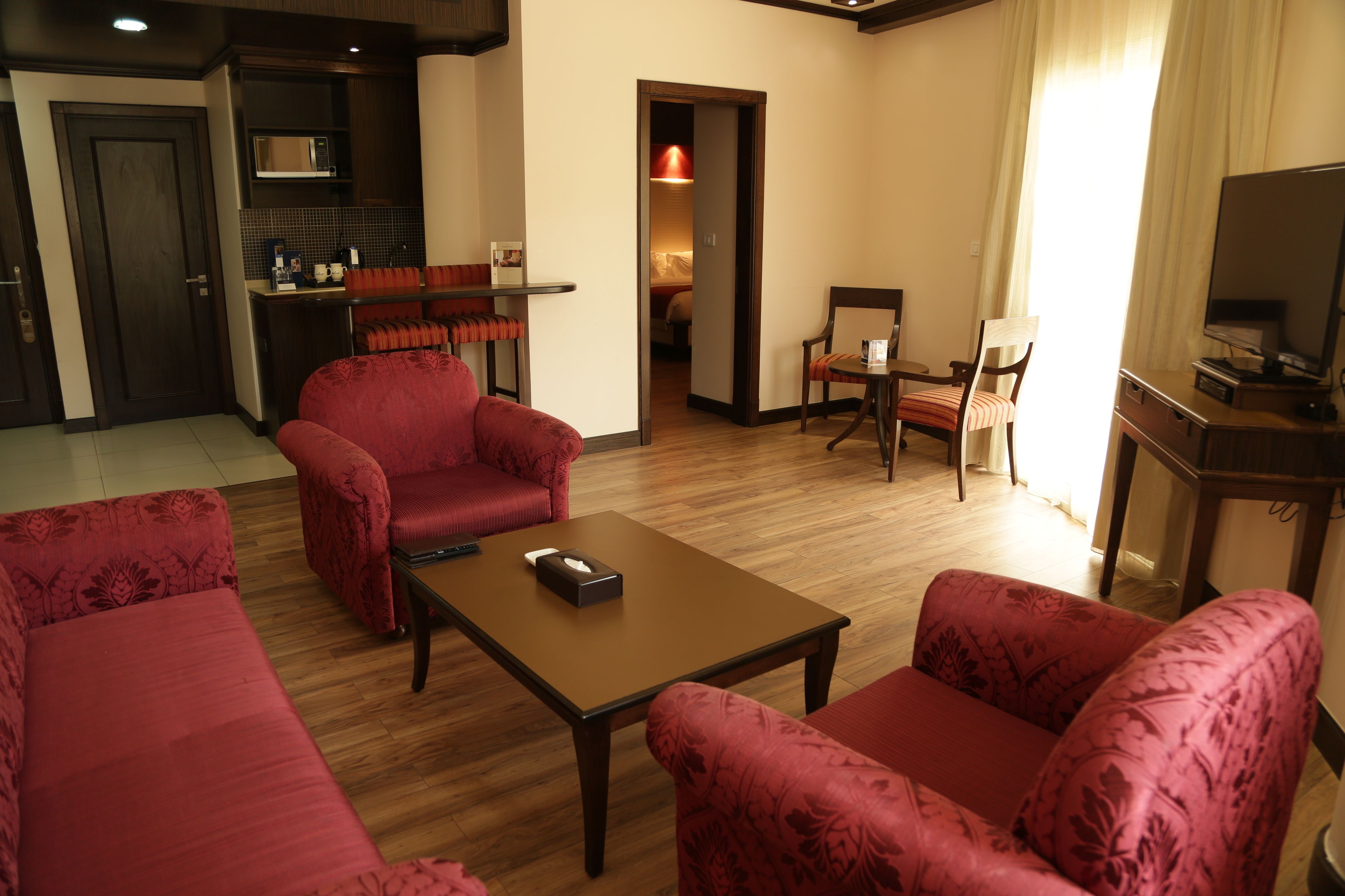 Habitación de hotel con piso de madera, sofás tapizados en rojo, una pequeña mesa, un televisor y una cocina con microondas.