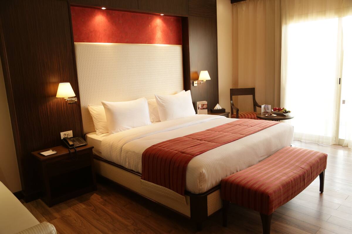 A hotel room with a large bed, wooden headboard, bedside lamps, a small table with a fruit bowl, and a window with curtains.