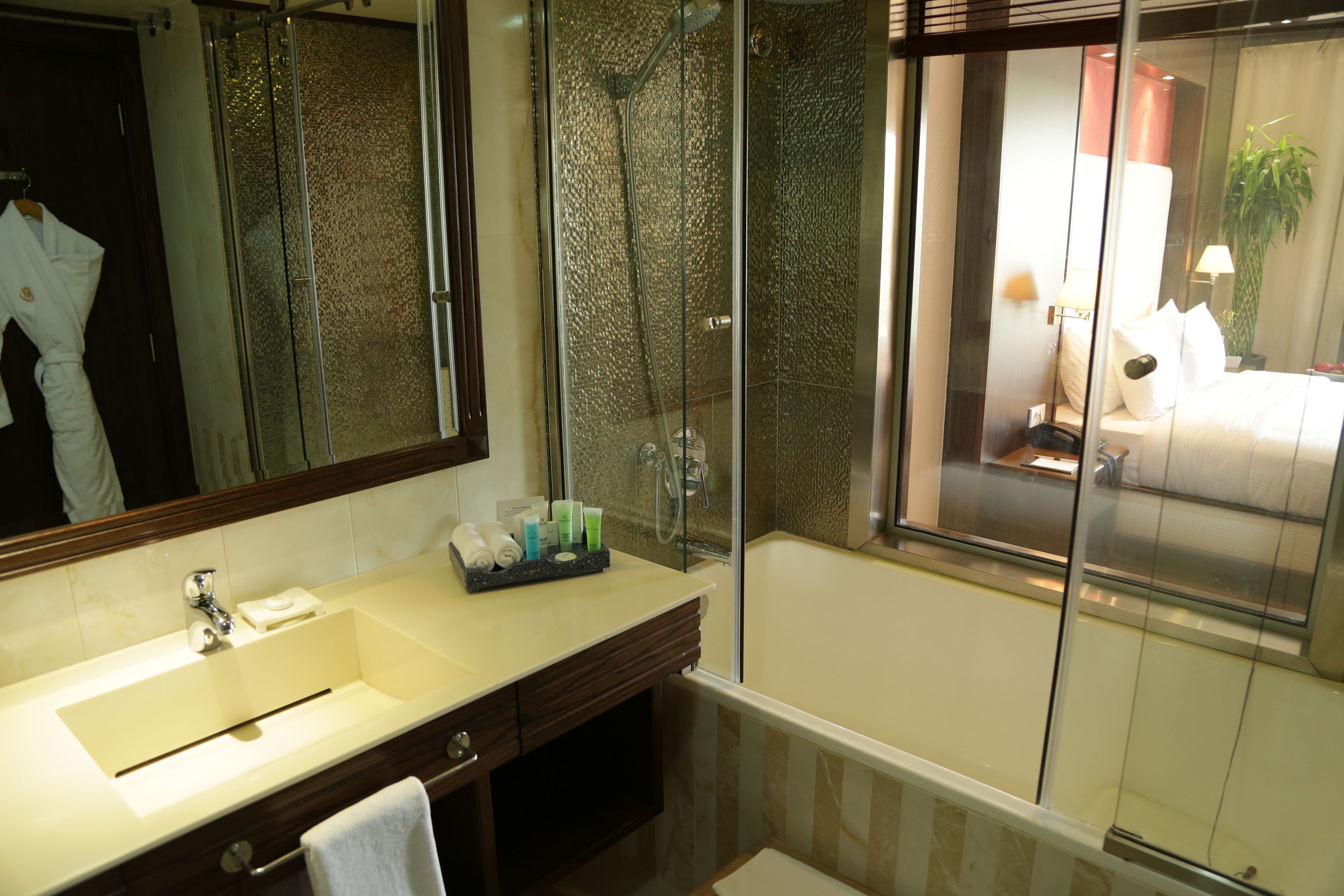 Un baño con un espejo grande, un lavamanos, una ducha y vista a un dormitorio a través de una puerta de vidrio.