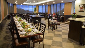 A spacious dining area with multiple tables set for meals, large windows offering a view, and a modern ceiling design. - Golden Lili Resort & Spa (Aley)