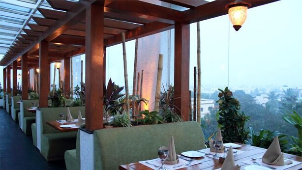 A restaurant with wooden tables and green cushioned seating, arranged in a covered outdoor area with large windows. - Aurick Boutique Hotel (Bengaluru)