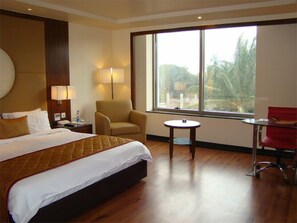 A hotel room with a large bed, a chair, a small table, and a window with a view of palm trees.