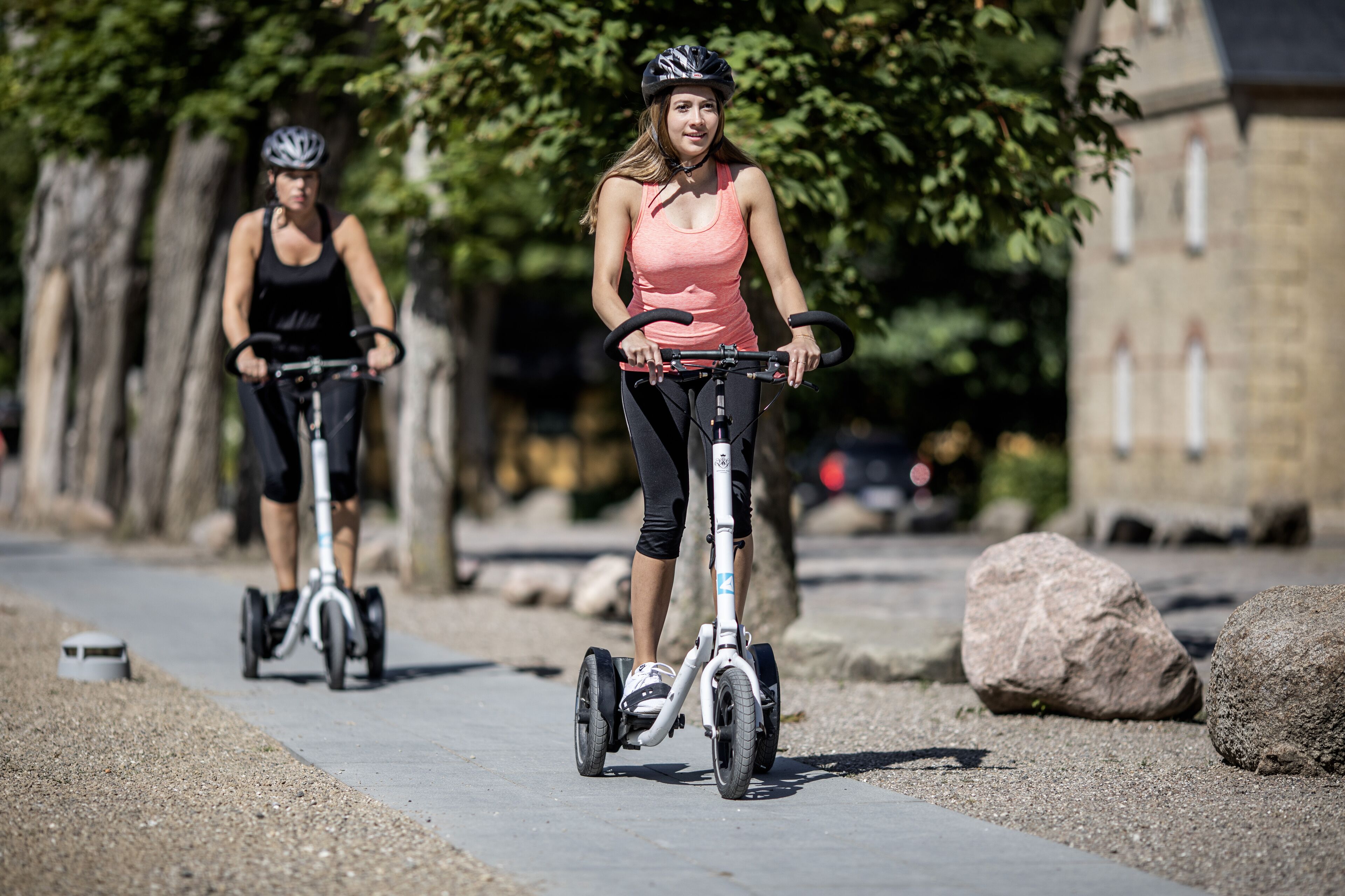 Two people riding electric scooters on a paved path with trees and a building in the background.