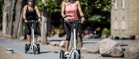 Two people riding electric scooters on a paved path with trees and a building in the background.