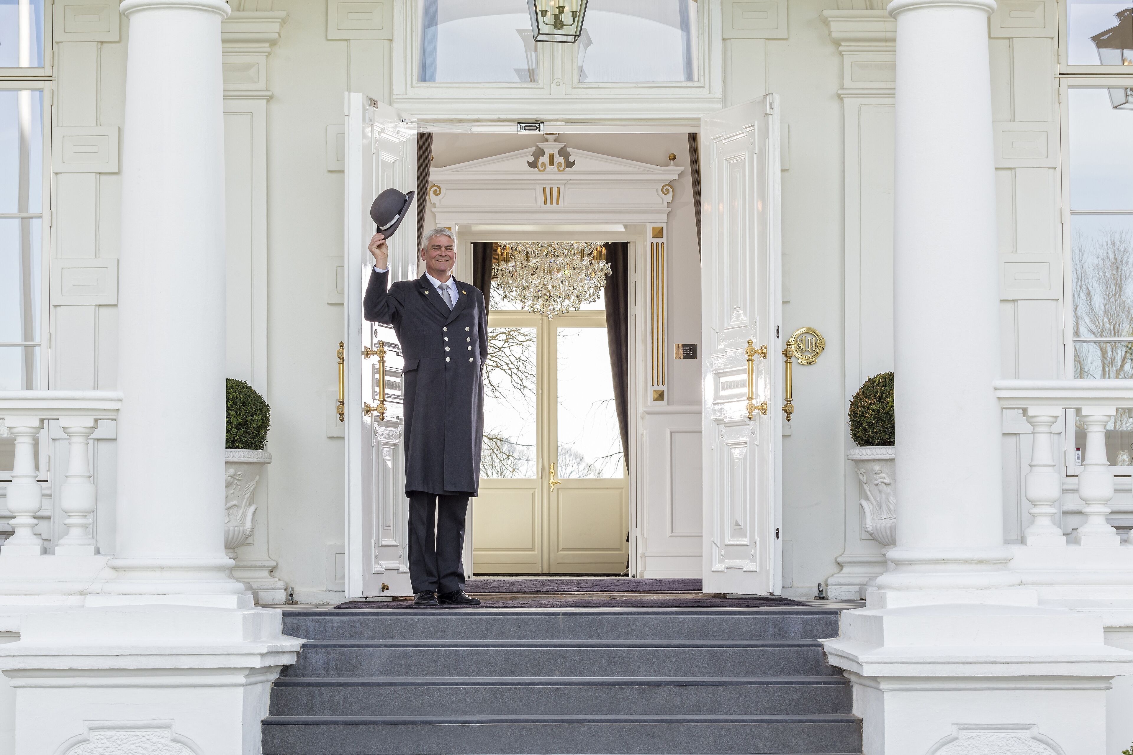 A grand entrance with a man in a suit standing on the steps, holding a hat.