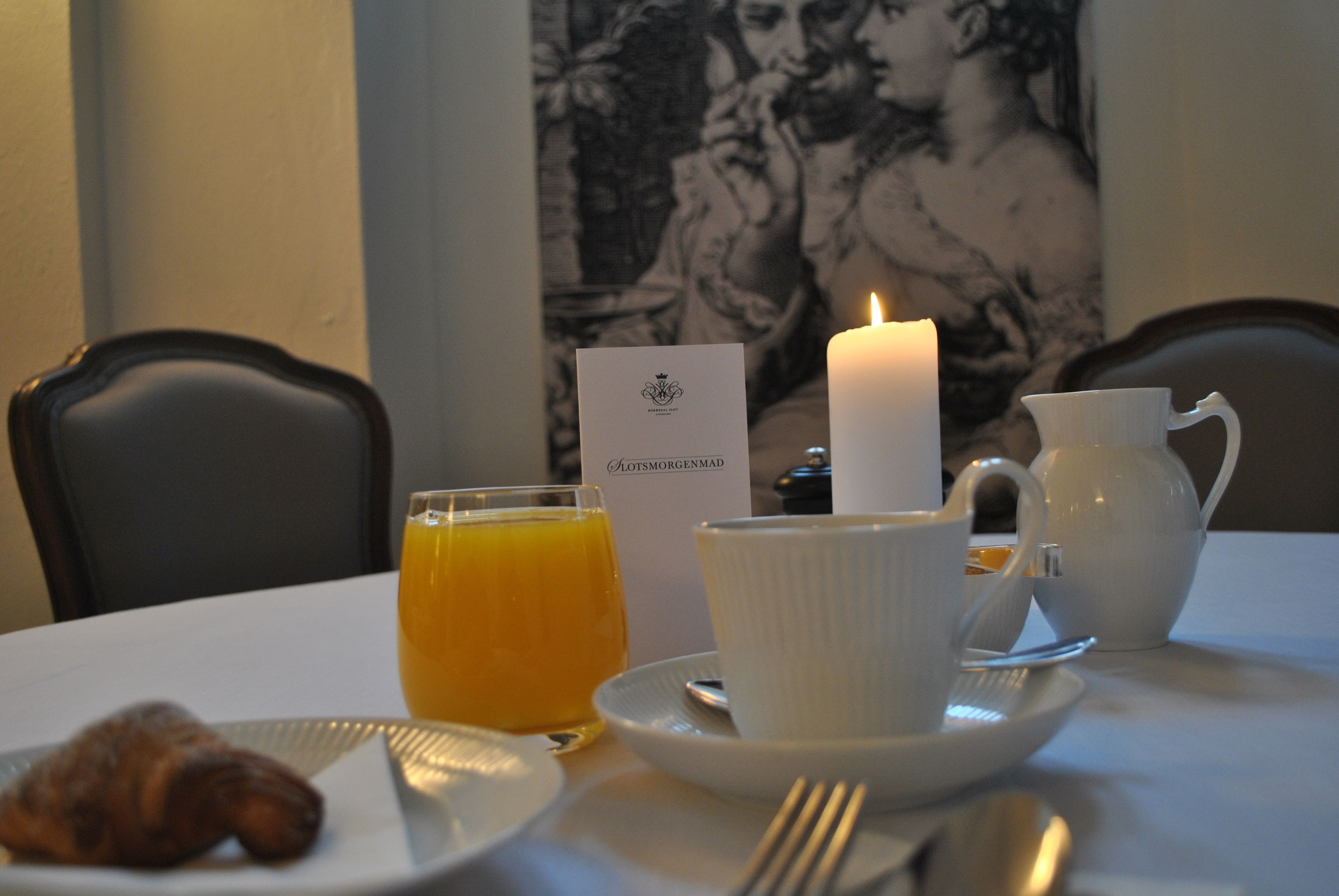 Um café da manhã com um croissant, suco de laranja, uma bule de chá branco e uma vela sobre uma mesa.