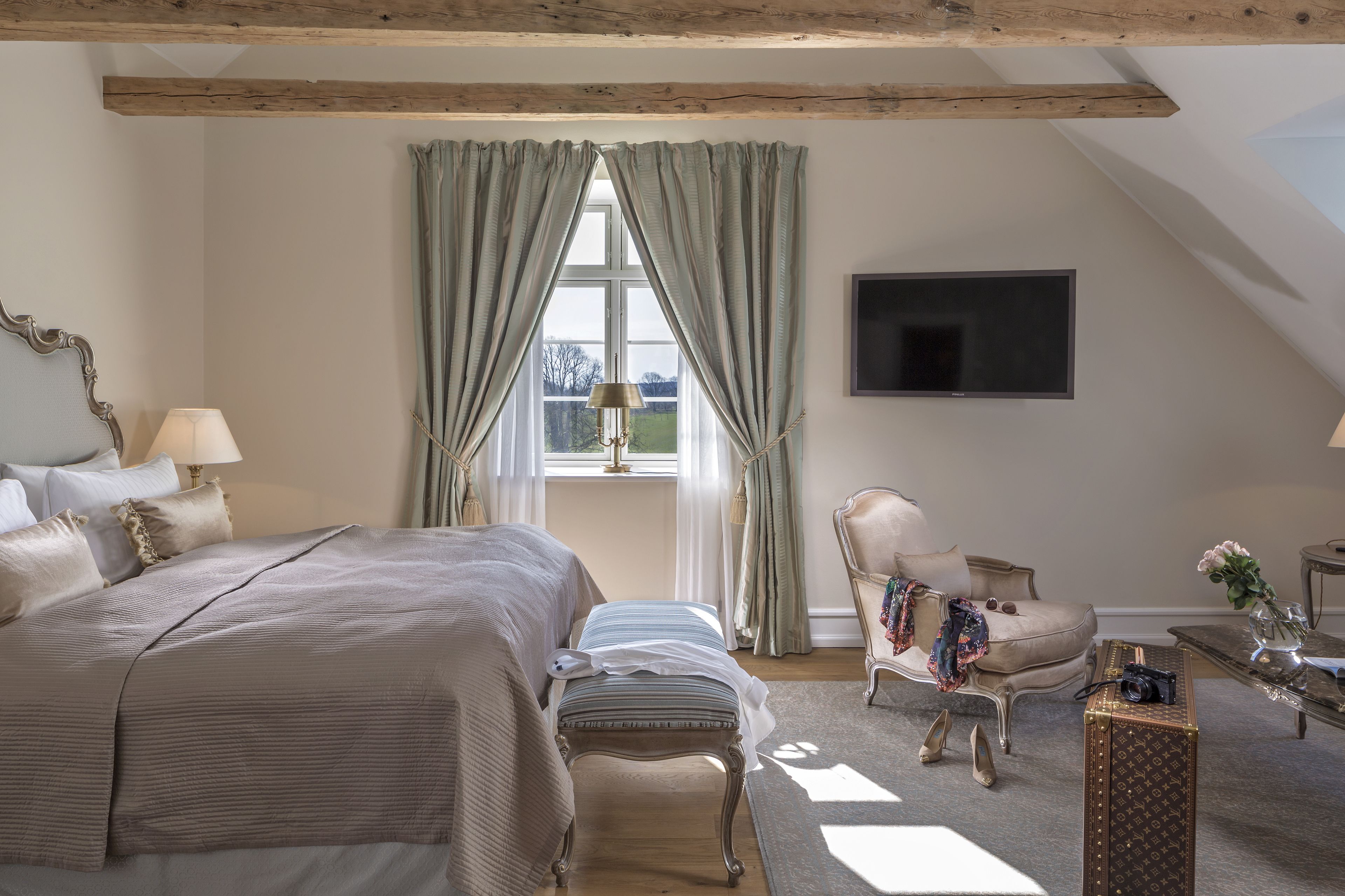 A bedroom with a large bed, a chair, a small table, and a TV mounted on the wall.