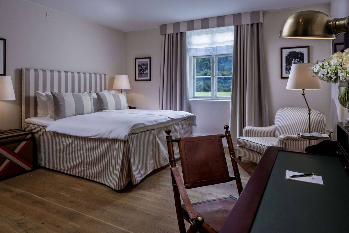 Habitació d'hotel amb un llit gran, una cadira de fusta, un escriptori i una finestra amb cortines.