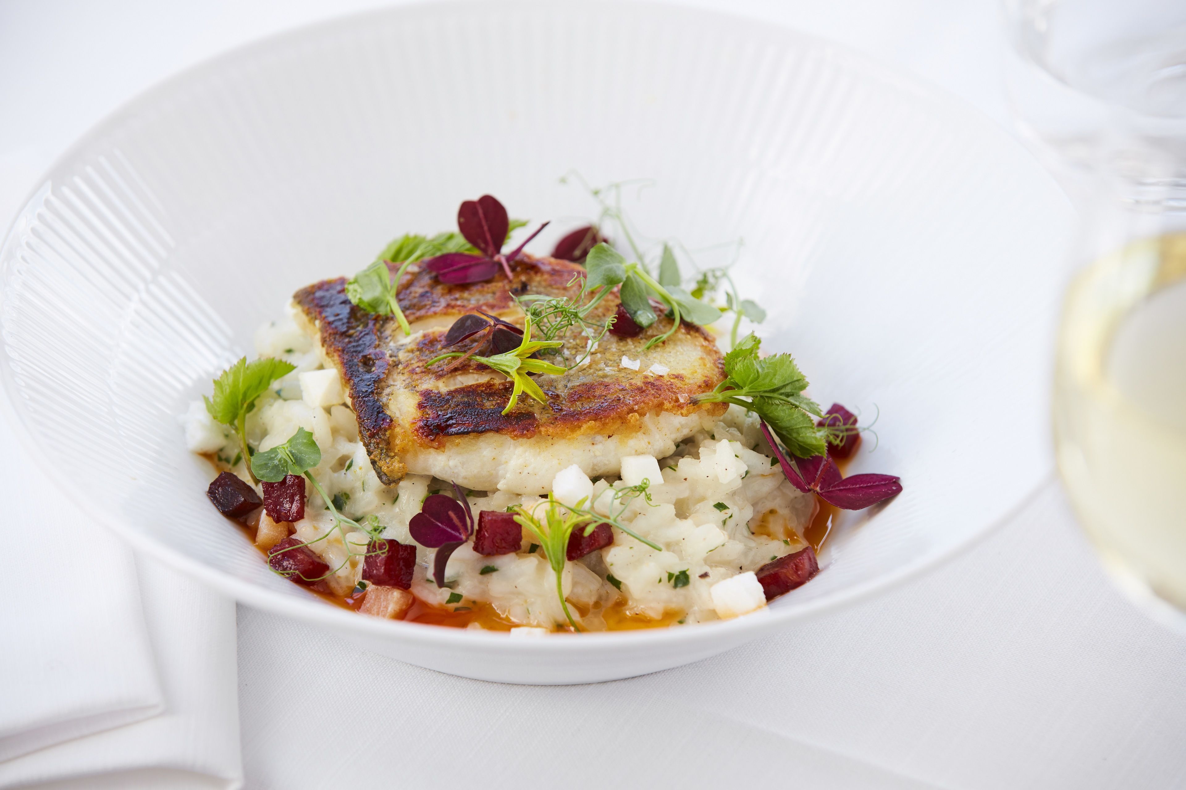 Une assiette blanche contenant un morceau de poisson grillé, agrémenté d’herbes aromatiques et de jeunes pousses, servi sur un lit de riz et de légumes coupés en dés.