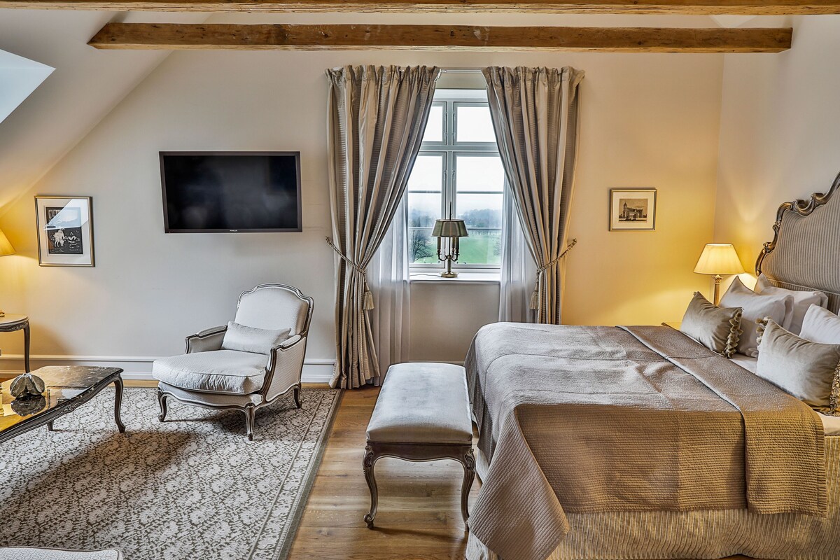 Un dormitorio con cama, una silla, una mesita, televisión y ventana con cortinas.