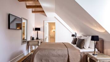 A bedroom with a sloped ceiling, a bed with a beige blanket, bedside tables with lamps, a mirror, and a wooden beam structure.