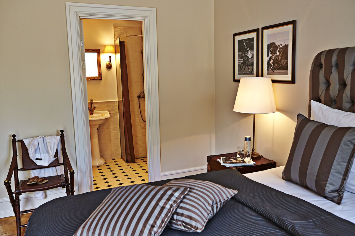 Habitación de hotel con cama, mesita de noche con lámpara, silla con una toalla y baño visible a través de una puerta abierta.