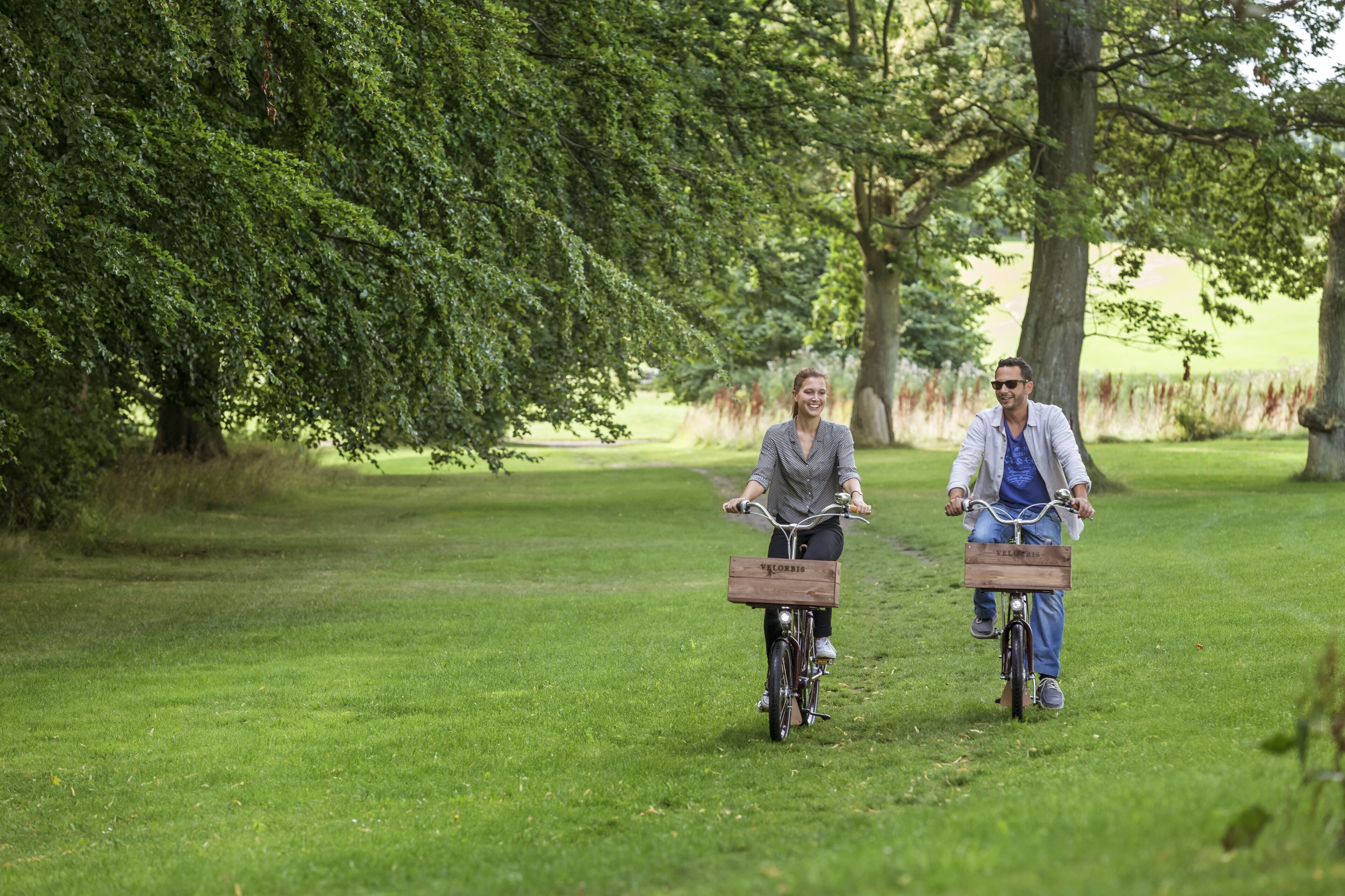 Två personer cyklar på en gräsbevuxen stig i en park.